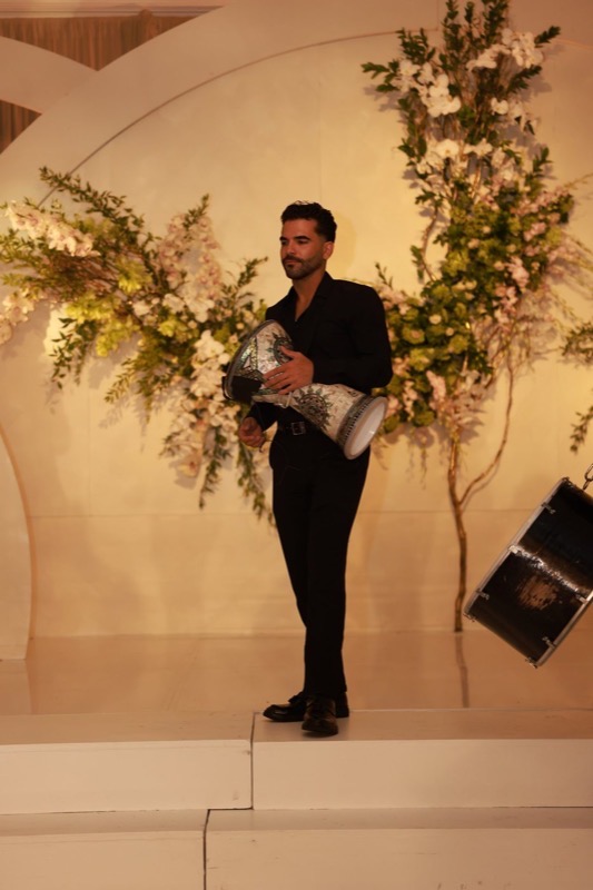 A Prestige Brothers derbake player performing solo against a floral stage backdrop